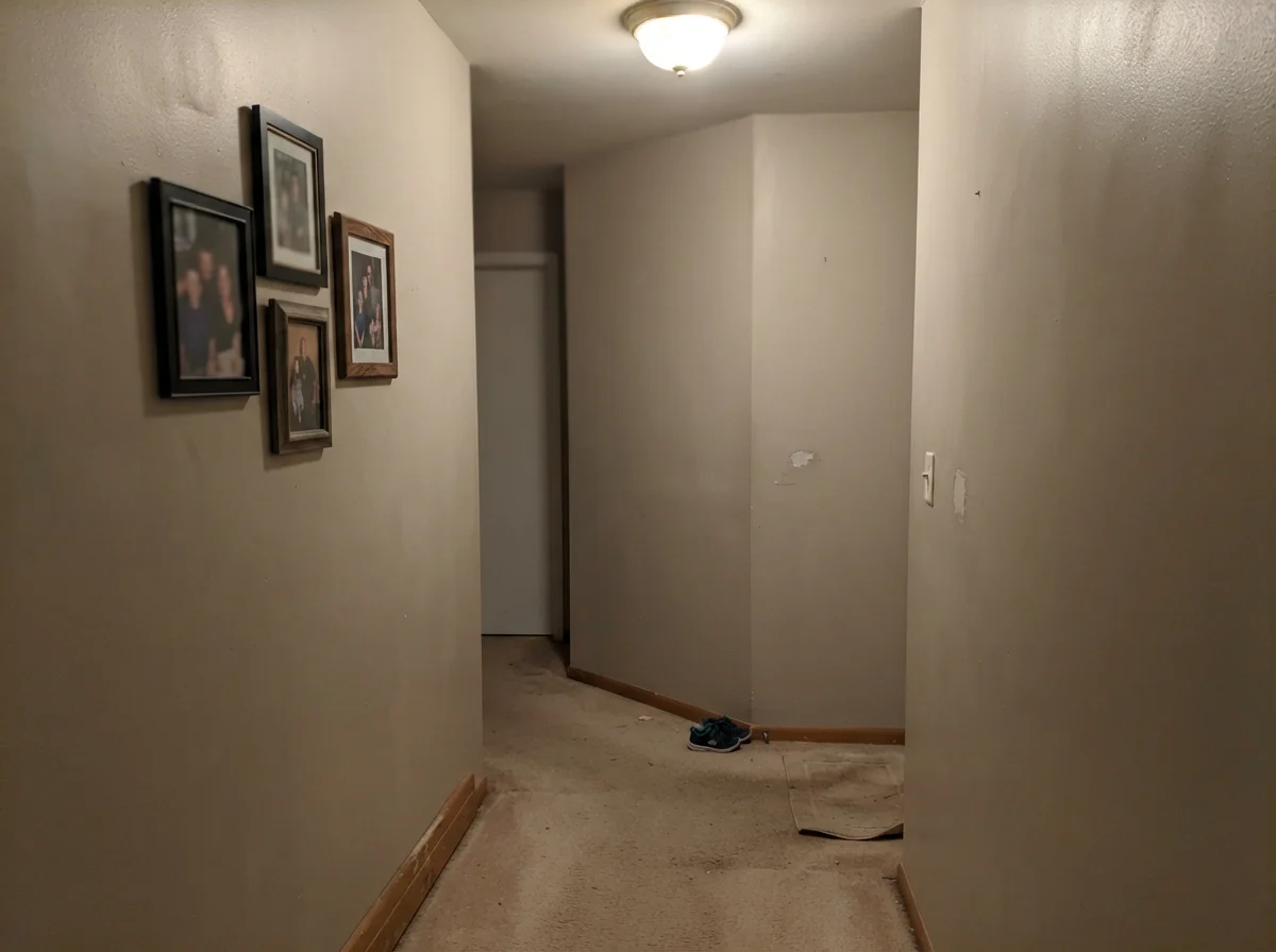 Dim windowless hallway corner where paint reads flat and muddy over beige carpet with framed photos.