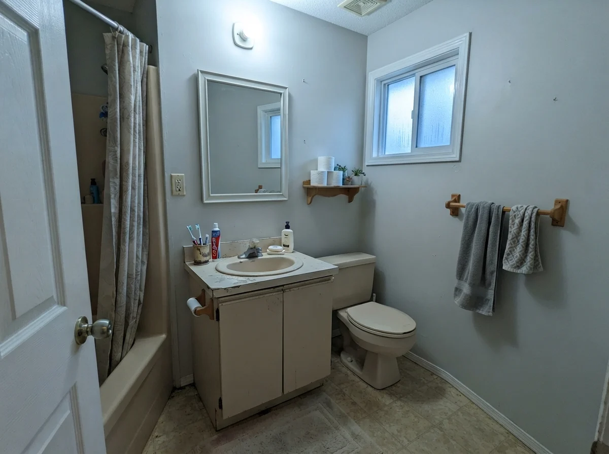 Dated bathroom showing cool cast on walls next to beige vanity on rainy day.